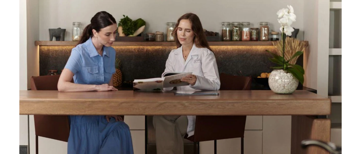 Two women sitting at a wooden table looking at a book, in front of a table full of jars