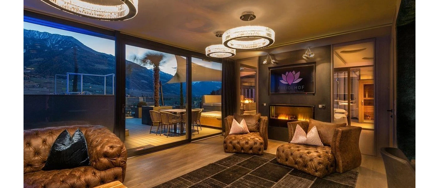 Cosy lounge area with glittering chandeliers, chesterfield-style leather armchairs in a brown shade, a wall-hung TV and floor-to-ceiling windows overlooking a terrace with mountain views