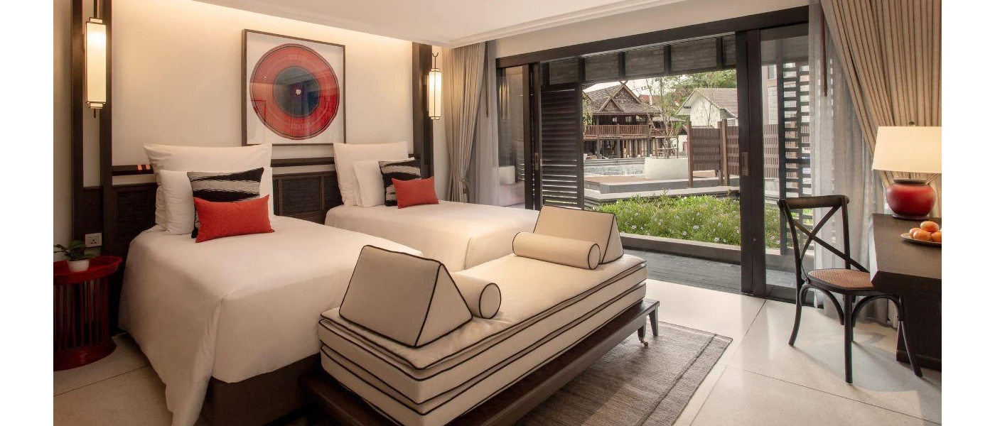 Hotel room with twin beds with white bedding, red and black cushions, dark-wood accents and a balcony door open to pool and garden views