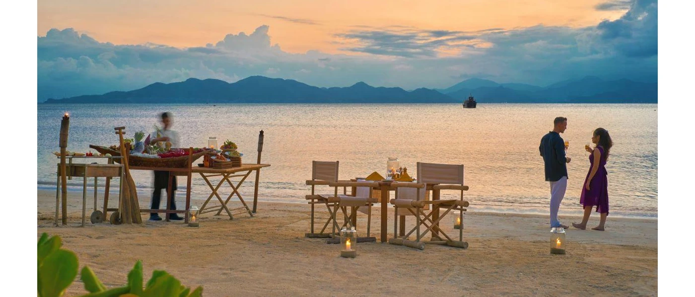 Beachfront at sunset: a couple enjoy a drink by the shoreline with a table set up for two behind them, and a staff member works behind a table full of food