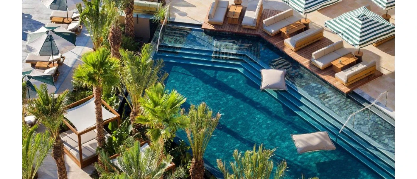 Corner swimming pool with steps leading into the water, lined with palm trees and terraces with white-cushioned loungers, sofas and striped parasols