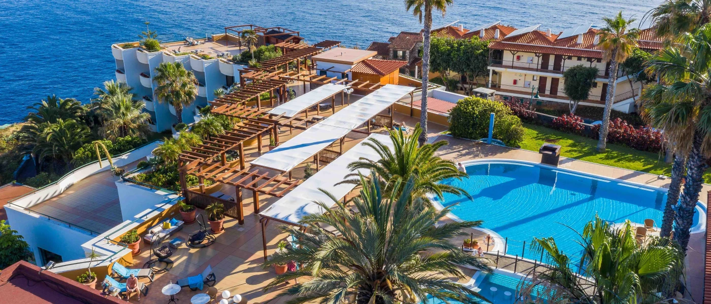 Aerial view of a hotel resort, with a swimming pool, palm trees, seating area, tall white buildings and the ocean in the background
