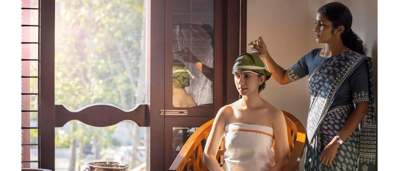 Indian lady in traditional dress pours oil onto the head of a guest in a white cloth with her hair wrapped in green
