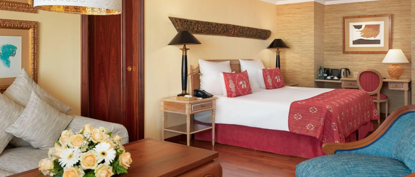 Hotel room with cream walls, white bedding, red soft furnishings, a blue chair and dark-wood furniture