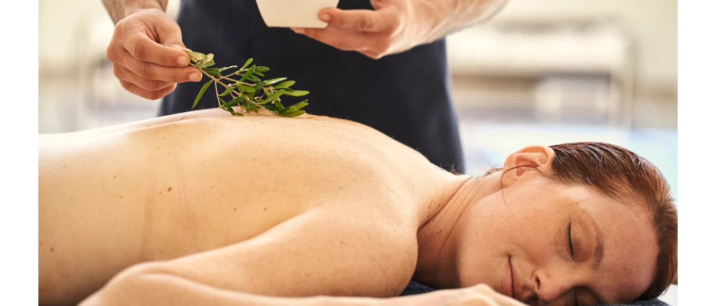 Smiling woman with eyes closed lies on her front, as a therapist caresses her back with a shrub