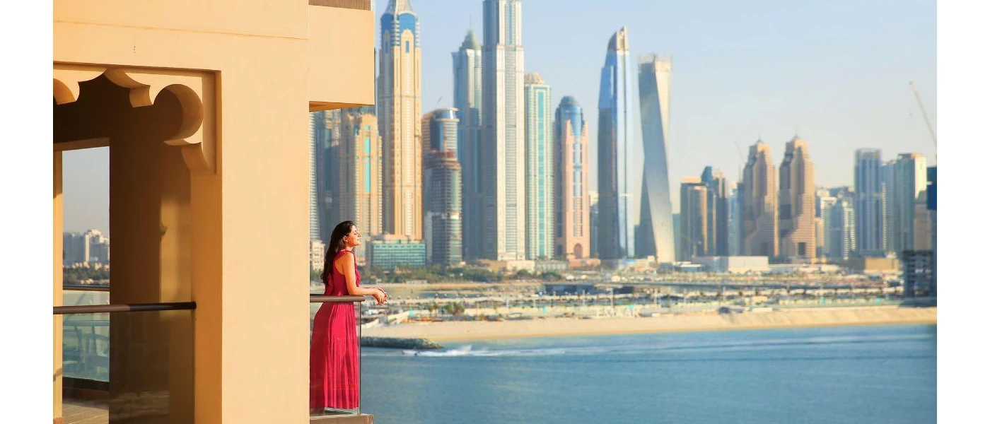 Woman in a pink dress gazes out at the Dubai skyline and sea from a glass-fronted balcony