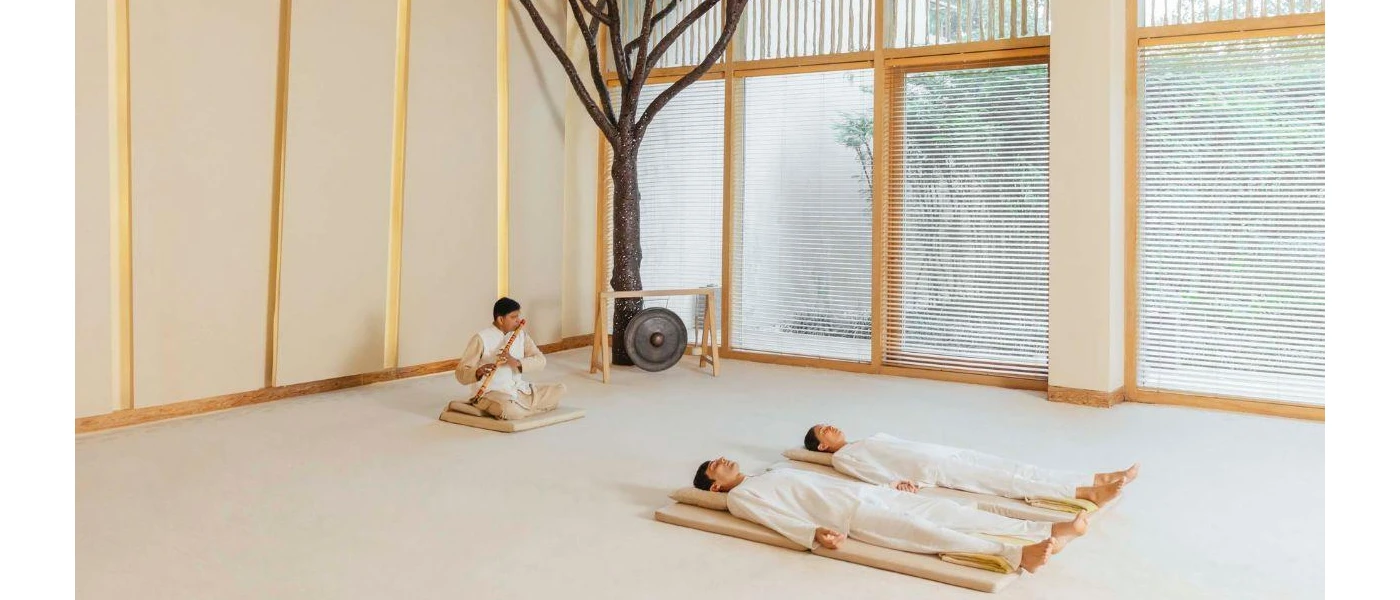 Man plays a flute-like instrument as two people lay with their eyes closed on mats in front of him in a bright studio space