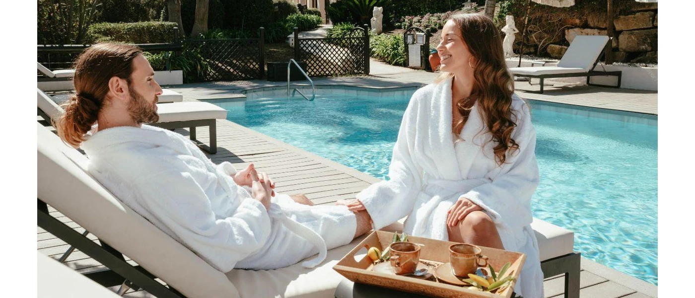 Smiling couple relax by the edge of a swimming pool in a sunny terrace, wearing white robes and enjoying a tray of herbal teas