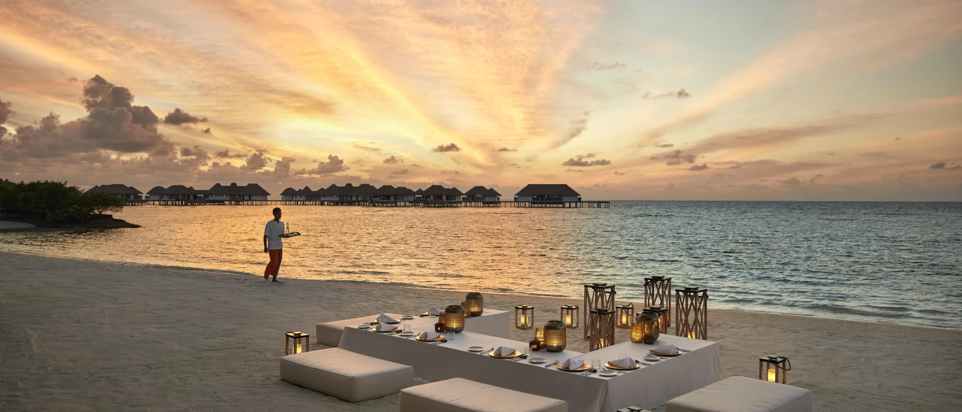 Low white L-shaped table laid up with a white tablecloth and candles, surrounded by cushioned benches on a sandy beach at sunset