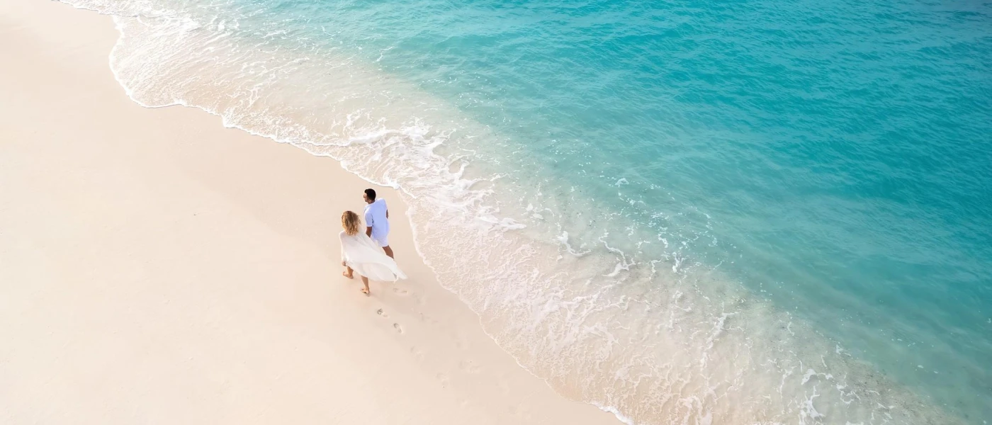 Couple in white strolling hand in hand on a white-sand beach, with vivid turquoise waves lapping against the shore