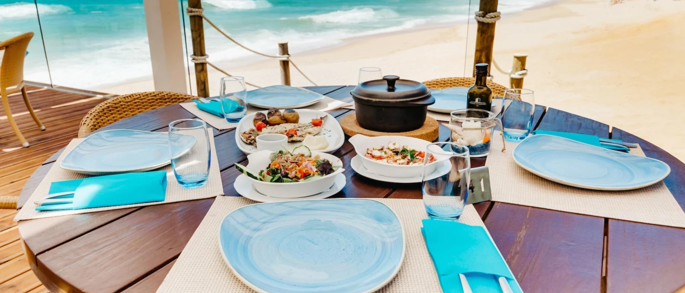 Round wooden table on the beachfront with blue plates and napkins and white plates of seafood and salad