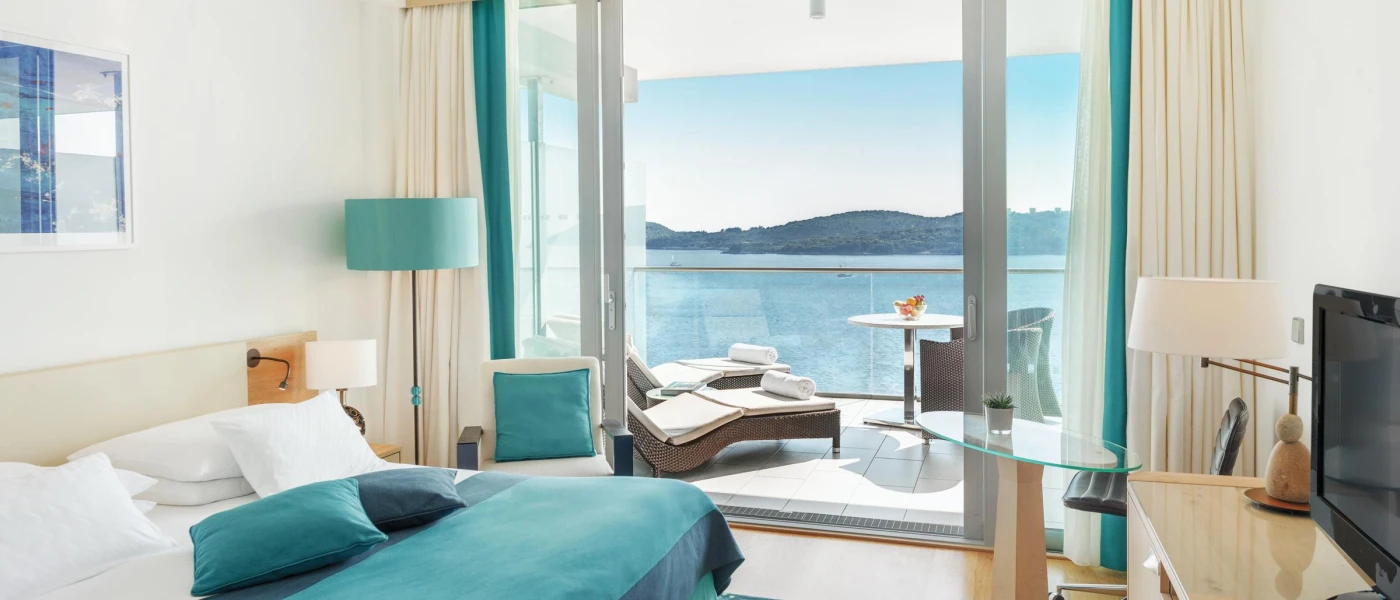 Modern hotel bedroom with white walls and bedding, blue soft furnishings, a desk with a TV and a furnished balcony with a sea view