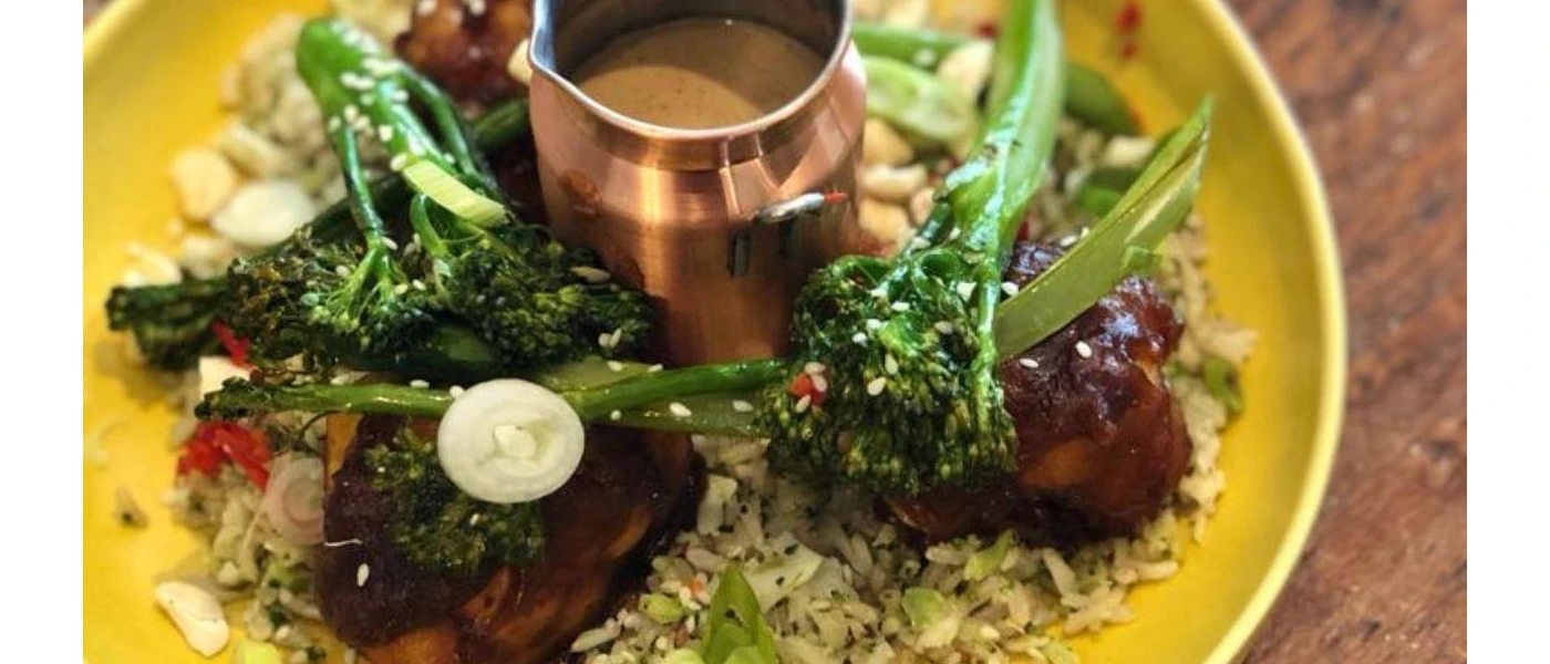 Yellow plate piled with vegetables and grains including tender stem broccoli, with a small jug of dressing at its centre