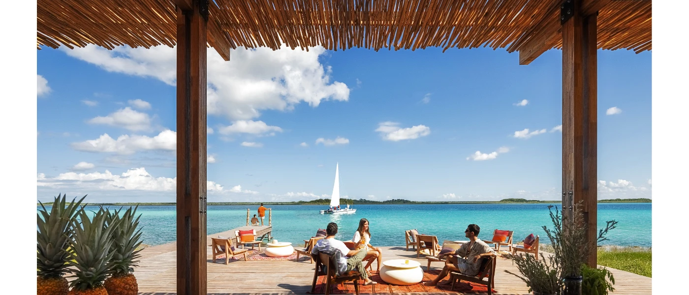 Thatched-roof pavilion framing a view of people sitting on chairs and beanbags on a waterfront deck under a blue sky