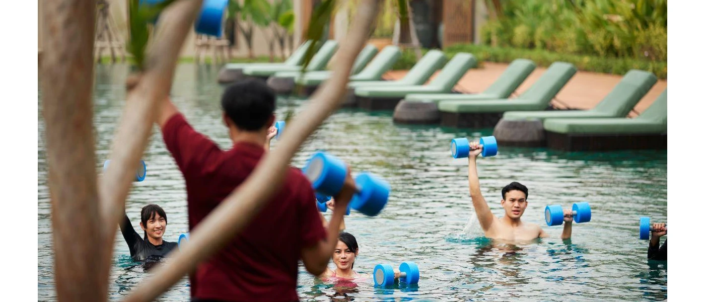 Staff member in red leads a class of people in a swimming pool holding up blue dumbbells