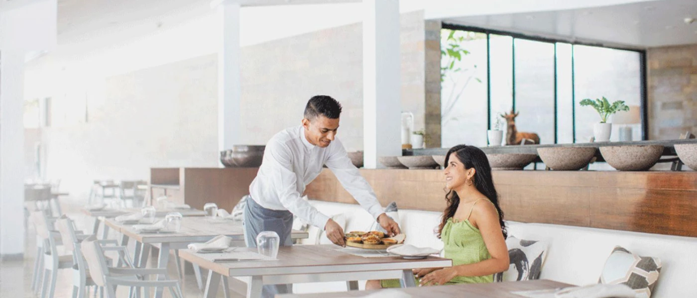 Waiter brings a smiling woman a plate of food in an airy restaurant with white walls, wooden furniture and large windows
