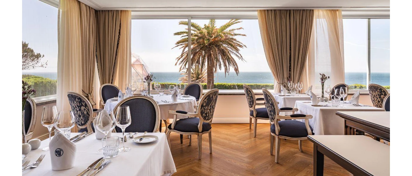 Bright restaurant with wooden floors, cream drapes and large windows with an ocean view, plus tables with crisp white tablecloths and elegant dark chairs