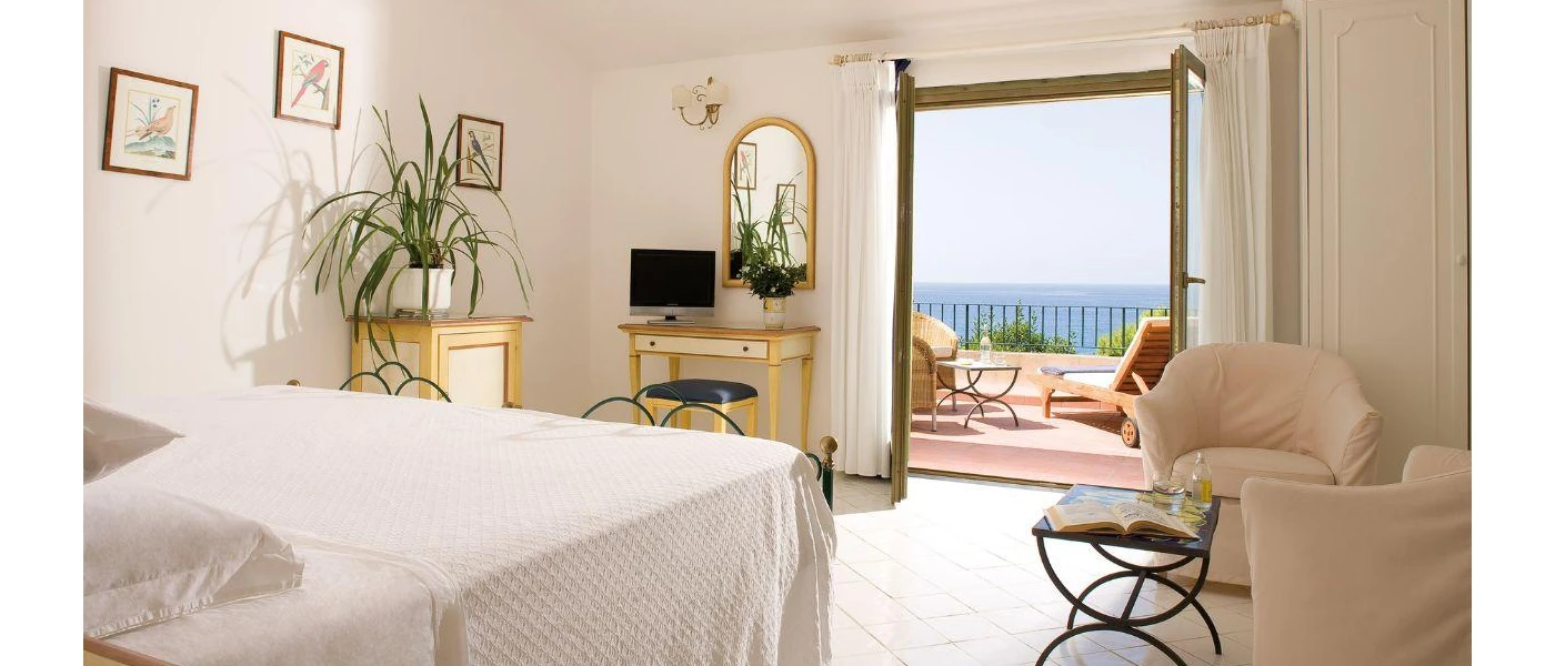 Airy bedroom with white bedding, potted green plants, a wooden dressing table and mirror, a TV and a balcony with a sea view