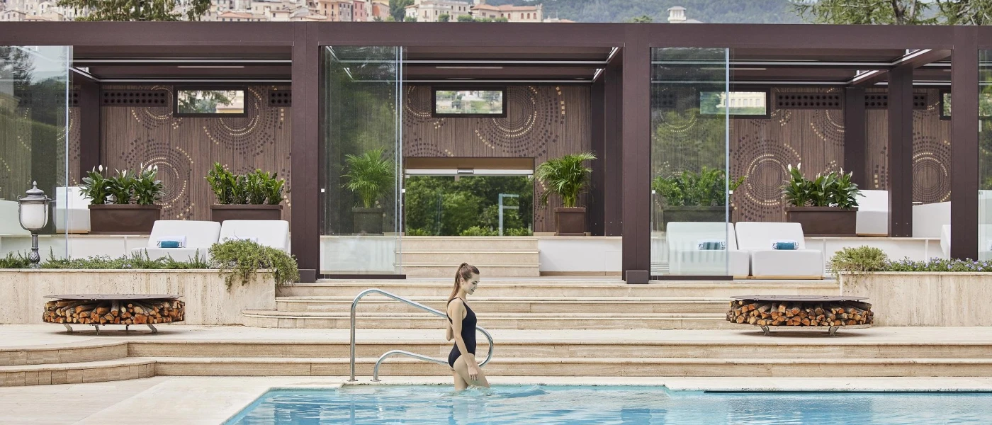 Woman in a black swimsuit walking into an outdoor pool with mountain peaks in the background