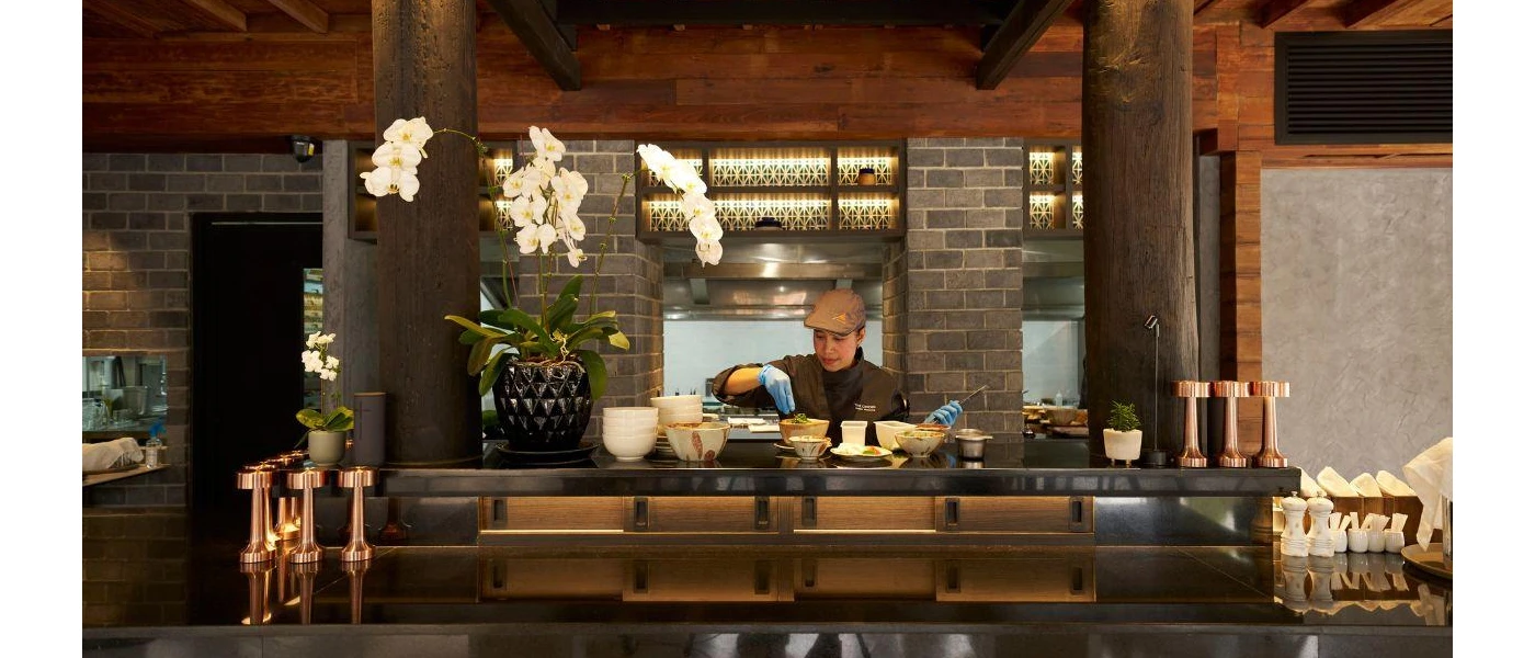 Female staff member in brown uniform and hat at a cooking station with a giant vase of orchids to one side