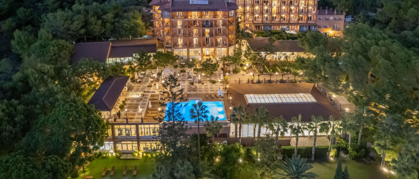 Multi-storey resort at night-time, as seen from the back with outdoor terraces, palm tree gardens and swimming pools 