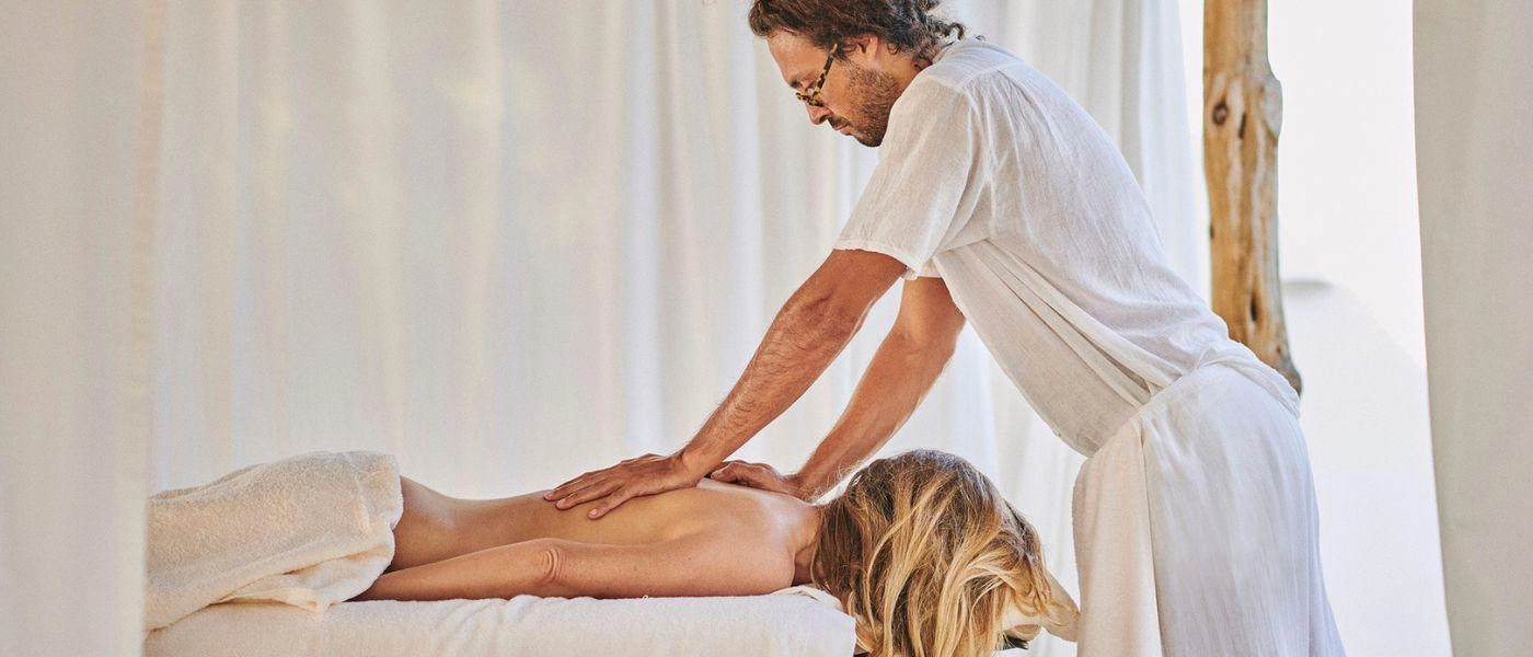 Male masseuse in white clothes works on a blonde female's back lying on a bed in a room with white curtains