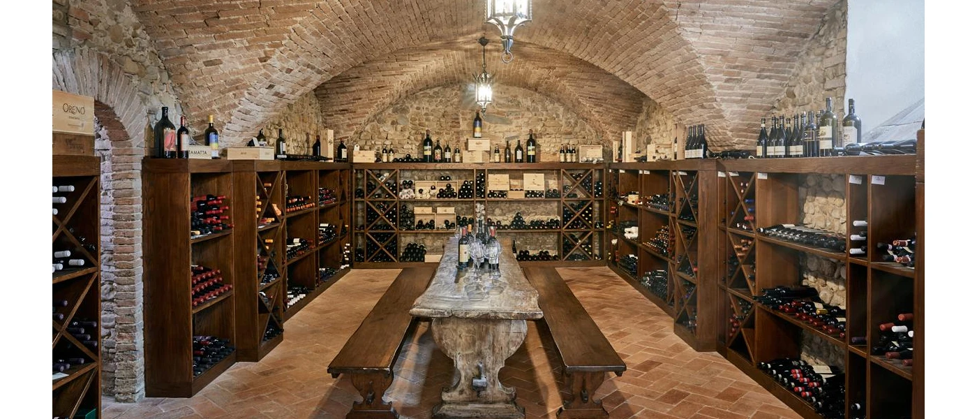 Medieval stone wine cellar with chunky wooden benches, a stone table and shelves stacked with bottles