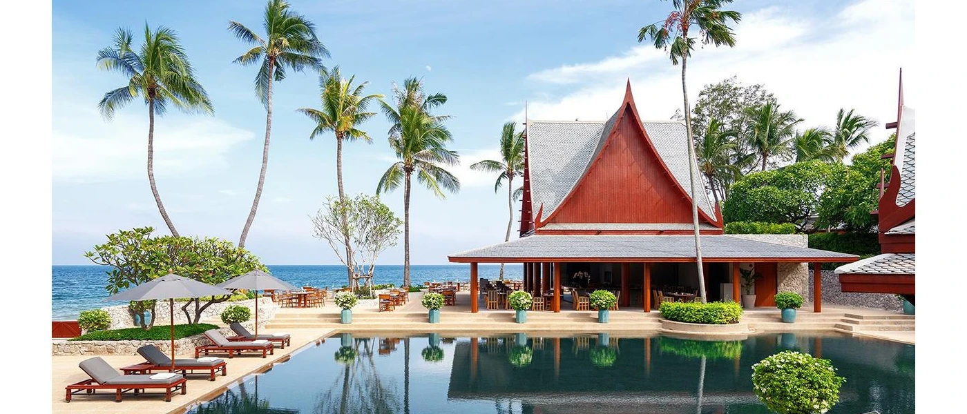 Swimming pool surrounded by wooden loungers and a temple-style building, with palm trees and the ocean in the background