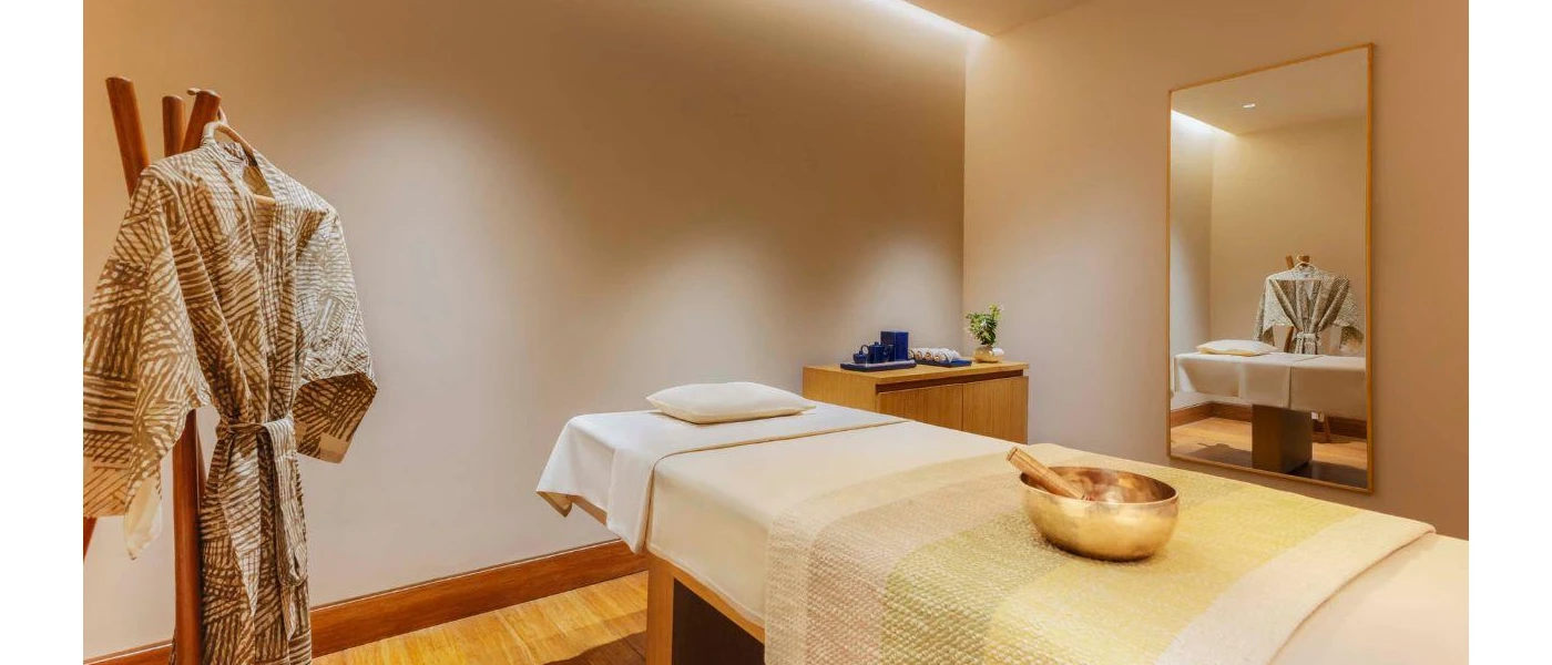 Treatment room with white walls, a massage bed with a sound bowl on top, a mirror, wooden vanity unit and a patterned robe hanging to one side