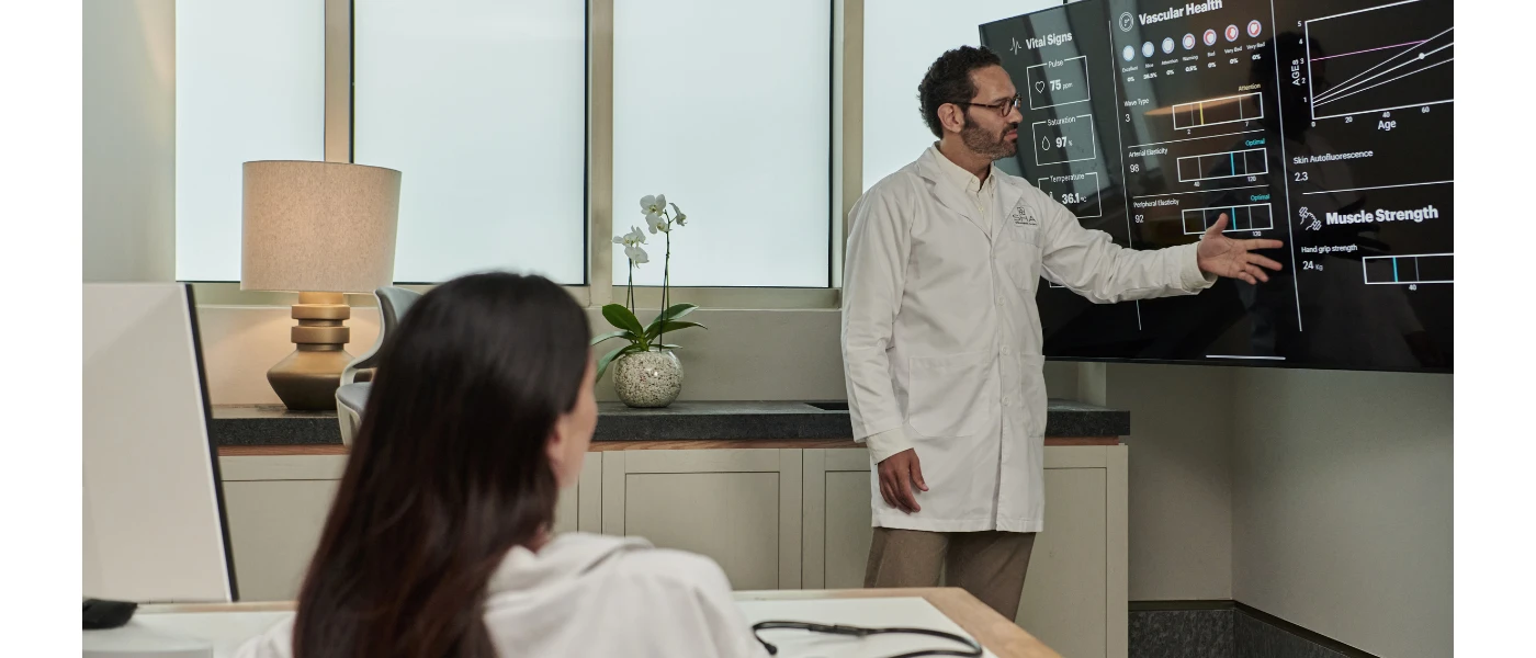 Health professional in a white coat gestures to a screen with readings on vascular health, vital signs and muscle strength, as a sitting woman looks on