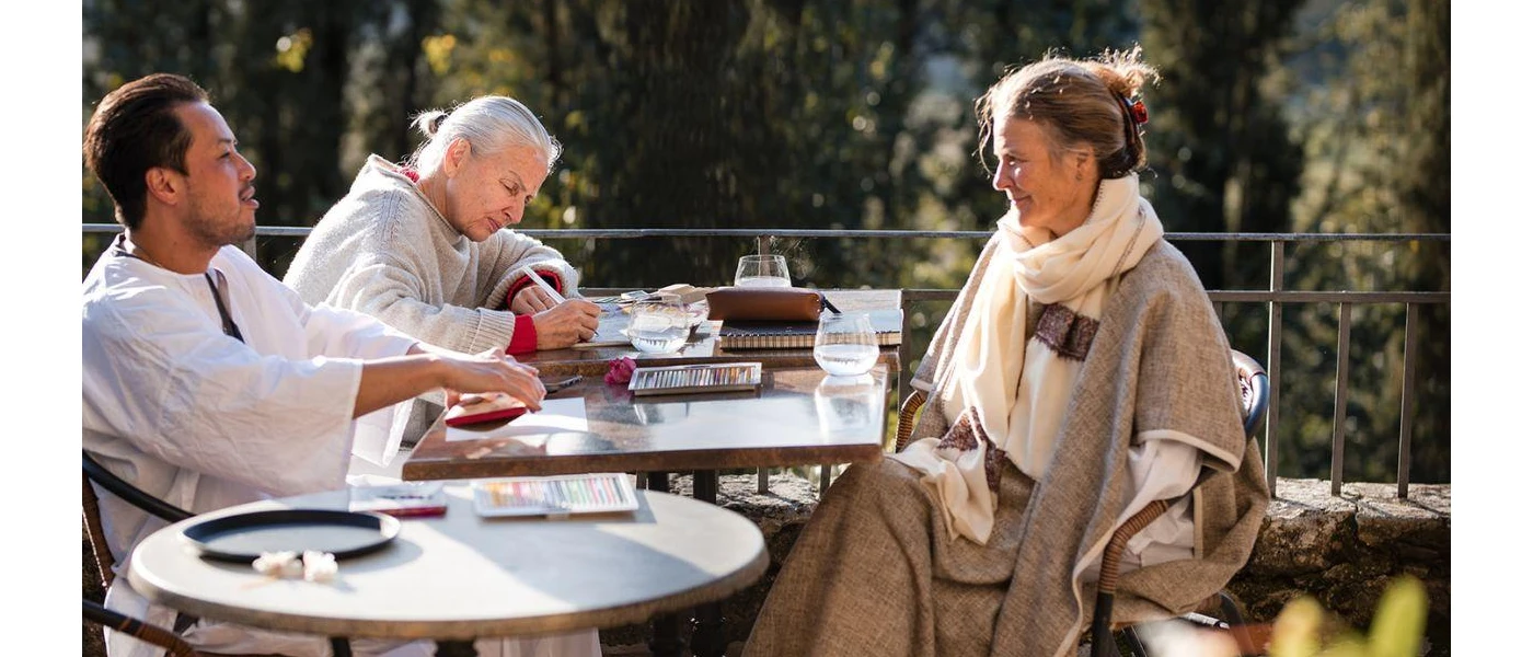 Group in loose fitting clothes sit around a table in the sunshine on a terrace with views of gardens