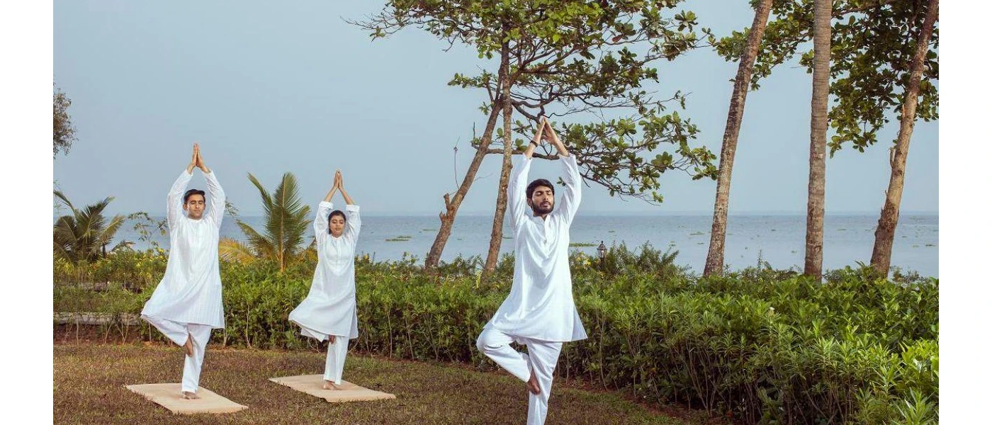 Three people in white Indian dress practice yoga outside in a waterfront garden 