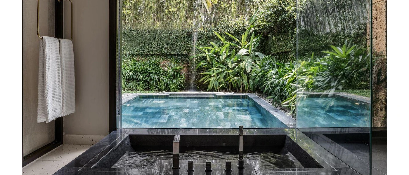 Bathroom with a bathtub and a huge window, overlooking a garden with a square swimming pool beyond the glass