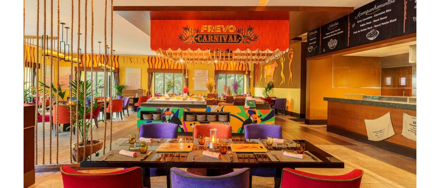 Colourful restaurant interior with yellow walls, purple and pink chairs, a station painted with toucans and a sign that reads 'Frevo Carnival'