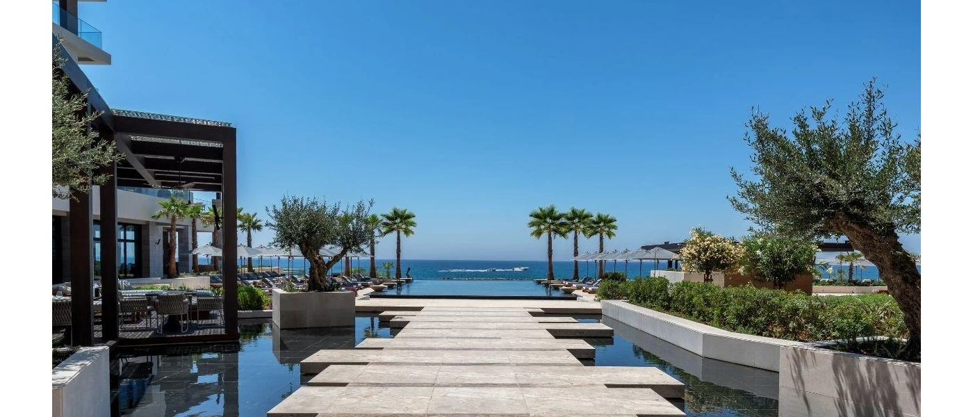 Sleek wooden decking over water leading to an infinity pool with a view of palm trees and the ocean