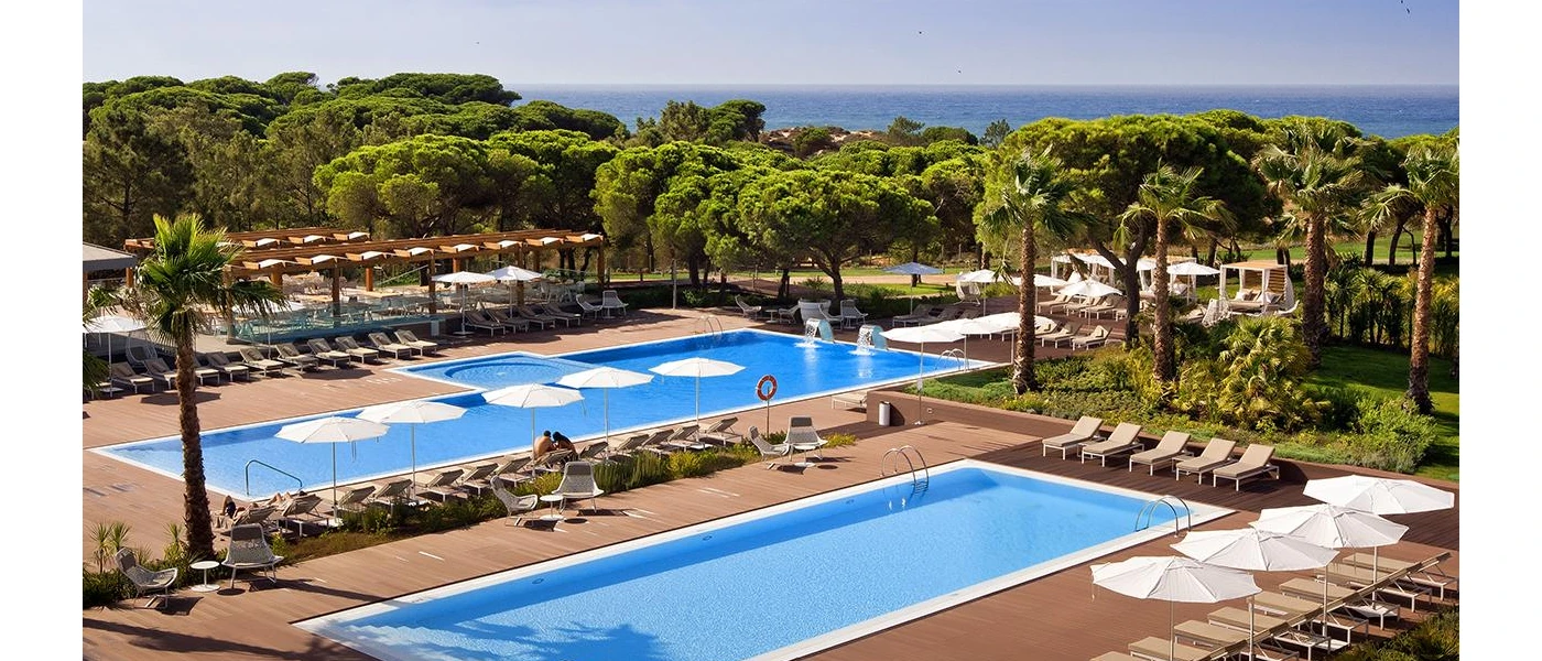 Two rectangular swimming pools in wooden terraces, surrounded by white parasols and loungers and landscaped gardens