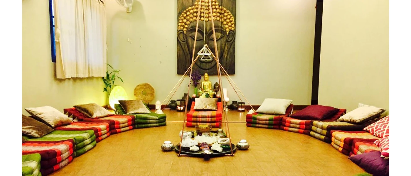 Room with brightly coloured cushions laid out in a circle, a large Buddha picture on the wall and a central set-up for some sort of ceremony