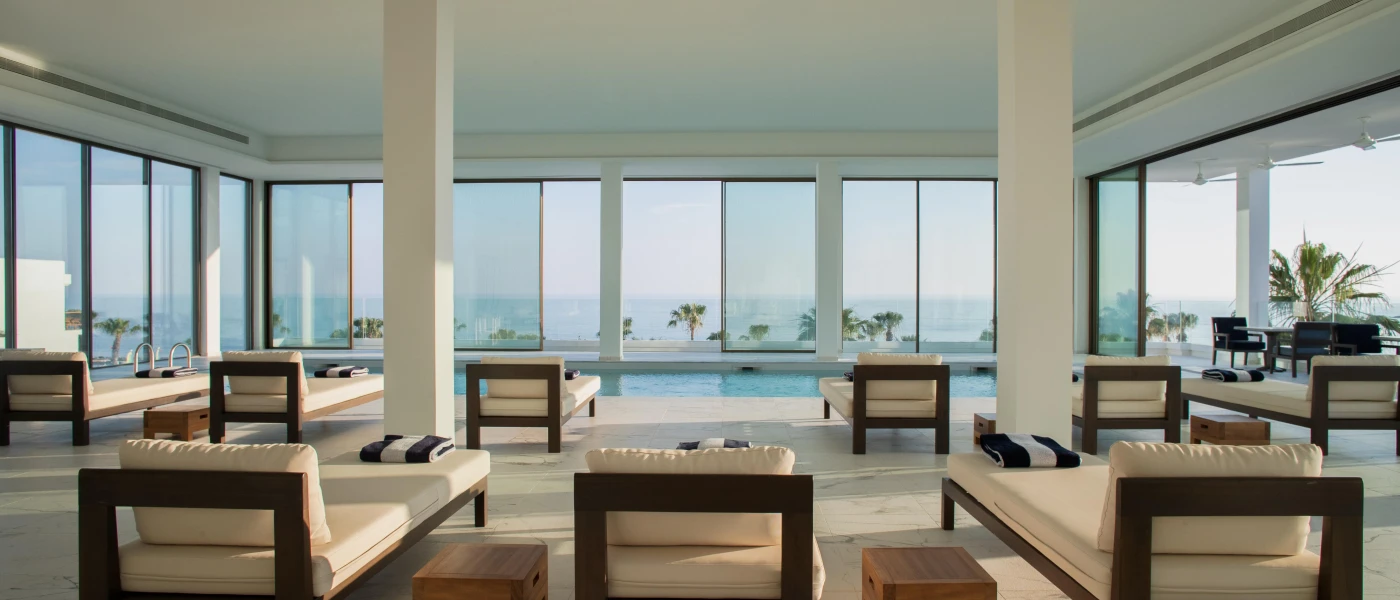 Indoor swimming pool surrounded by white pillars, white-cushioned dark-wood loungers with striped towels on them, and floor-to-ceiling windows with a sea view