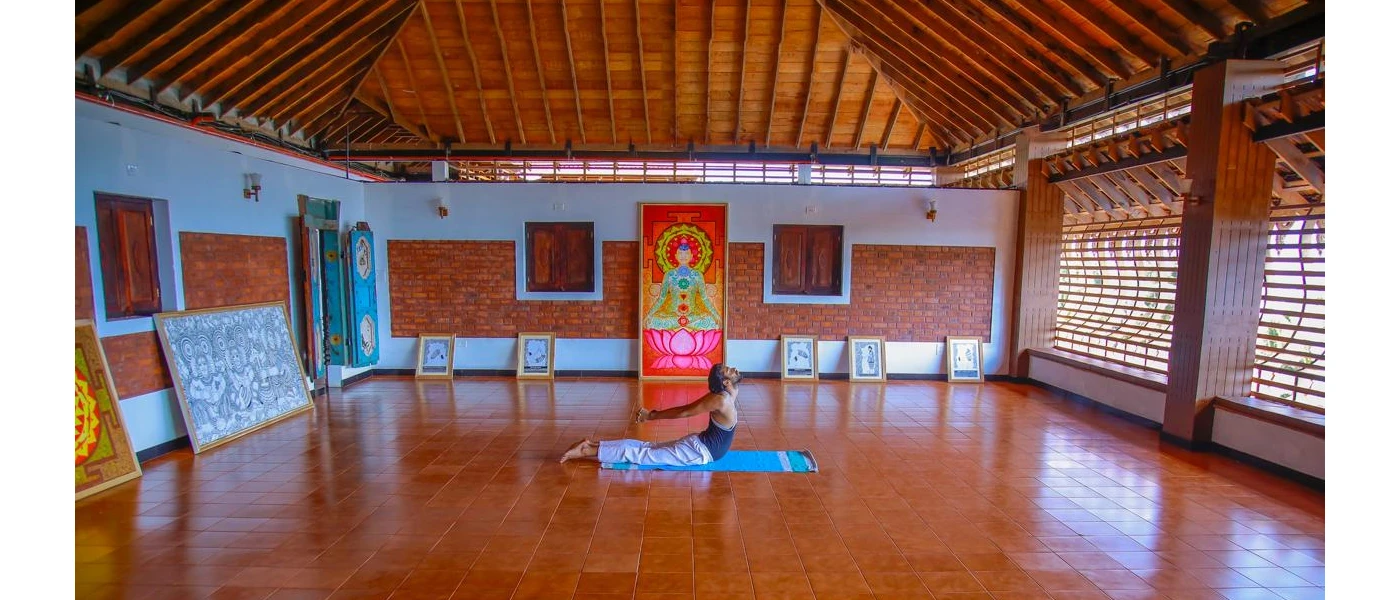 Man practices yoga on a blue towel in a studio with terracotta tiled floors, wood-beamed ceilings and colourful Hindu artworks