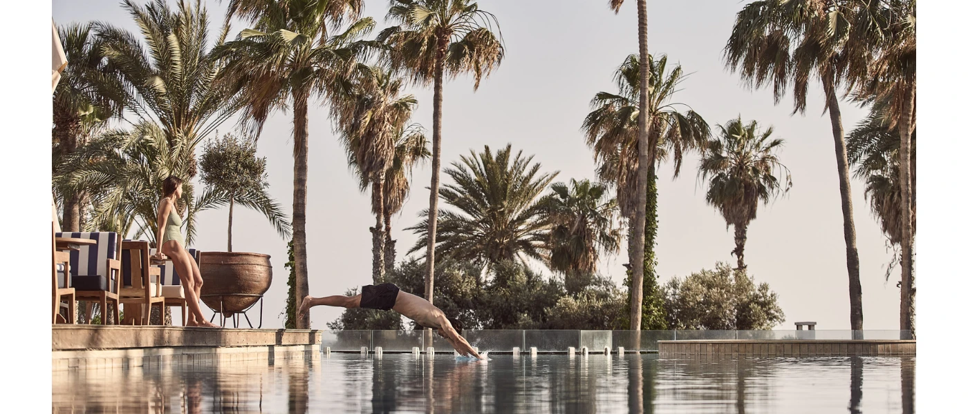 Man dives into a swimming pool edged by palm trees, as a woman in a lime swimsuit watches on