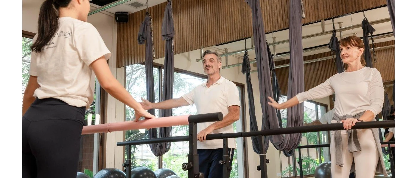 Man and woman in active wear stretch out while being supported by a bar as a staff member demonstrates in front of them