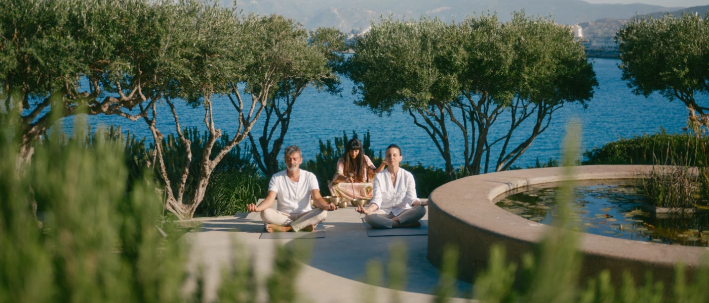 Three people in loose fitting clothes sit with their eyes closed and hands on their knees in a pretty garden with trees, a stone-walled pond and the ocean in the background