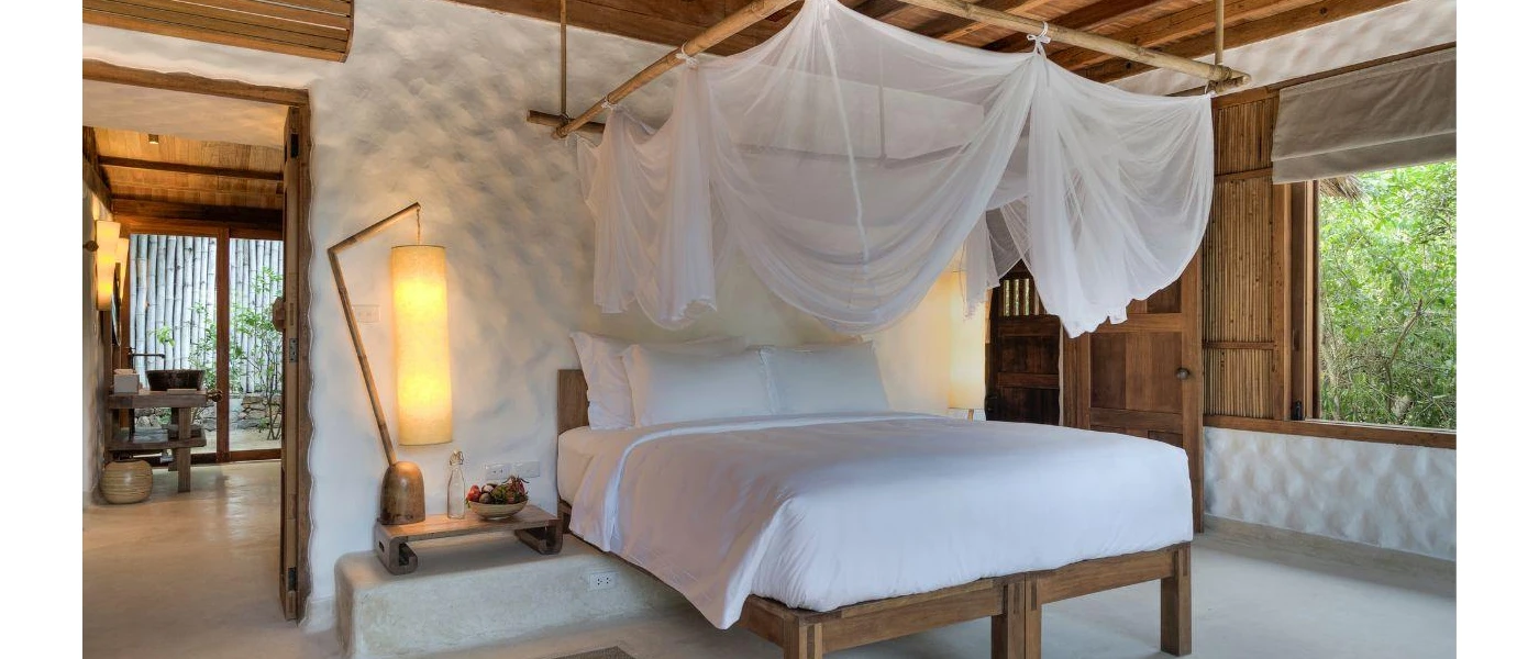 Villa bedroom with a wooden bed, wall panels and ceiling, a stone white wall, white bedding and sheer white drapes hung over the bed, plus lantern-style lighting 