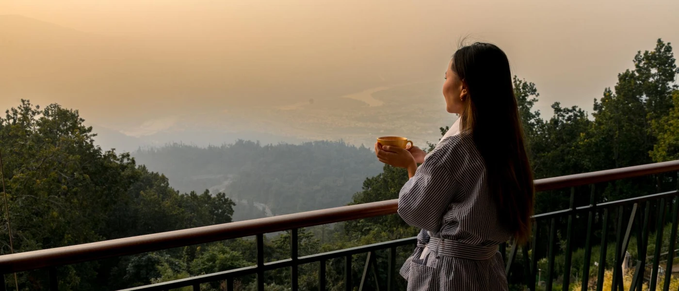 Woman in a white robe holding a yellow mug, overlooking the misty mountains as the sun rises in the distance 