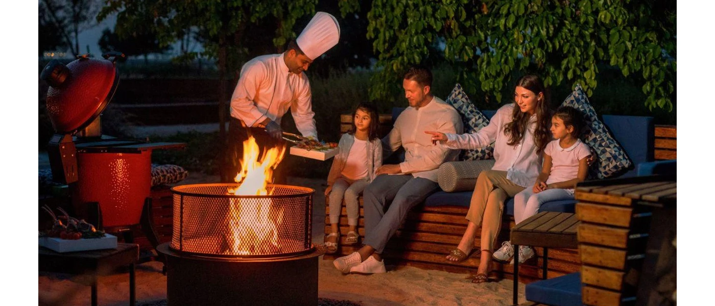 Man, woman and two daughters smile as they sit around a fire at night and a chef serves them from a barbecue