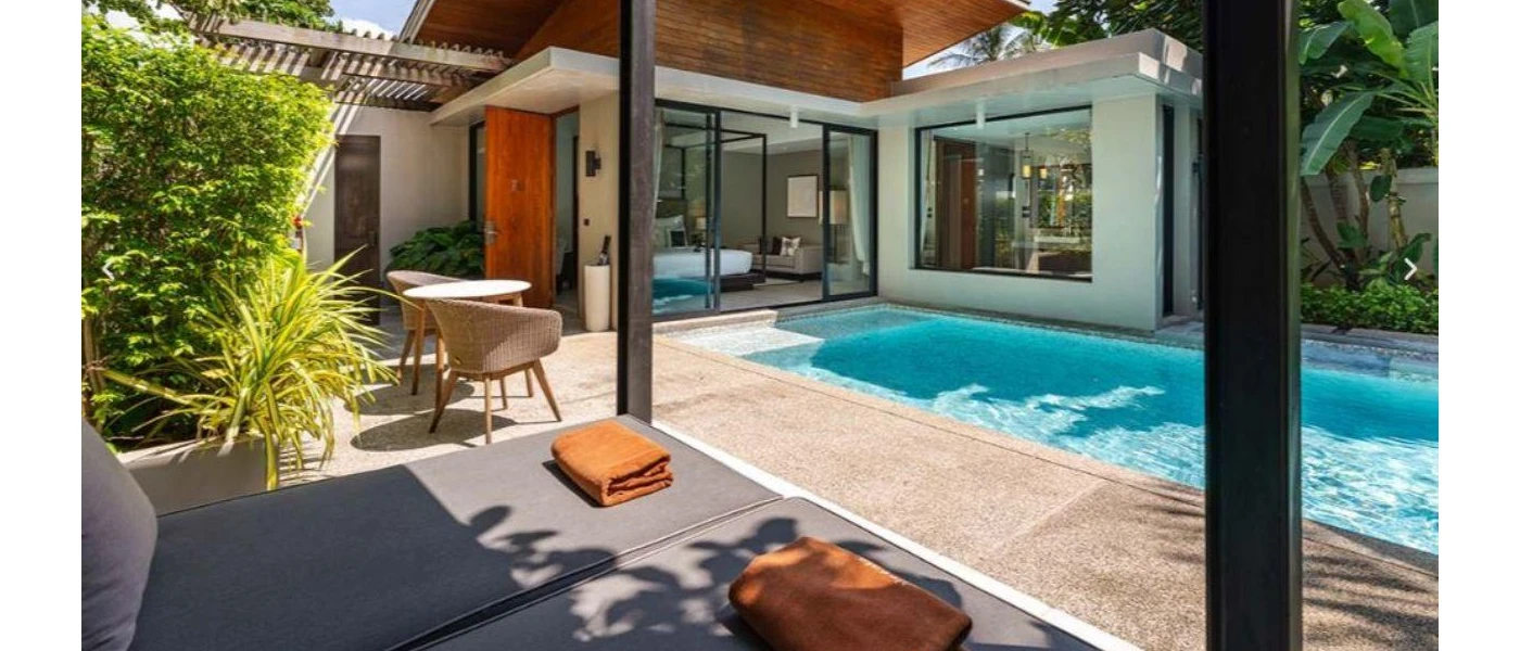 Swimming pool in a paved terrace with a a grey daybed and orange towels, plus palms and shrubs and a wooden table and chairs