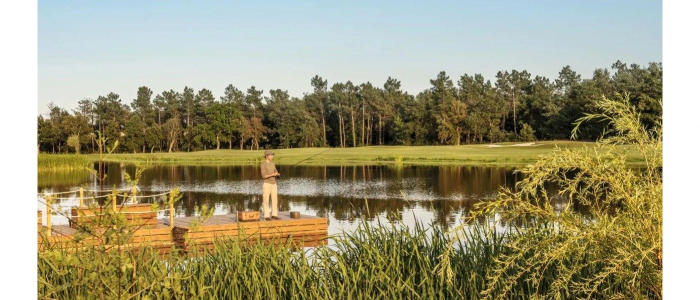 Man in camo attire fishes on a wooden jetty in a lake surrounded by lawns and woodland
