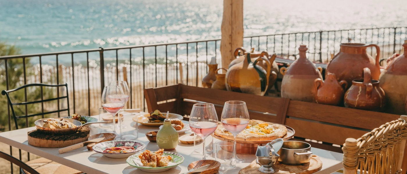 Table laid with rustic wooden bowls, glasses of rose, and a shelf full of pots next to a wrought-iron balcony with a sea view