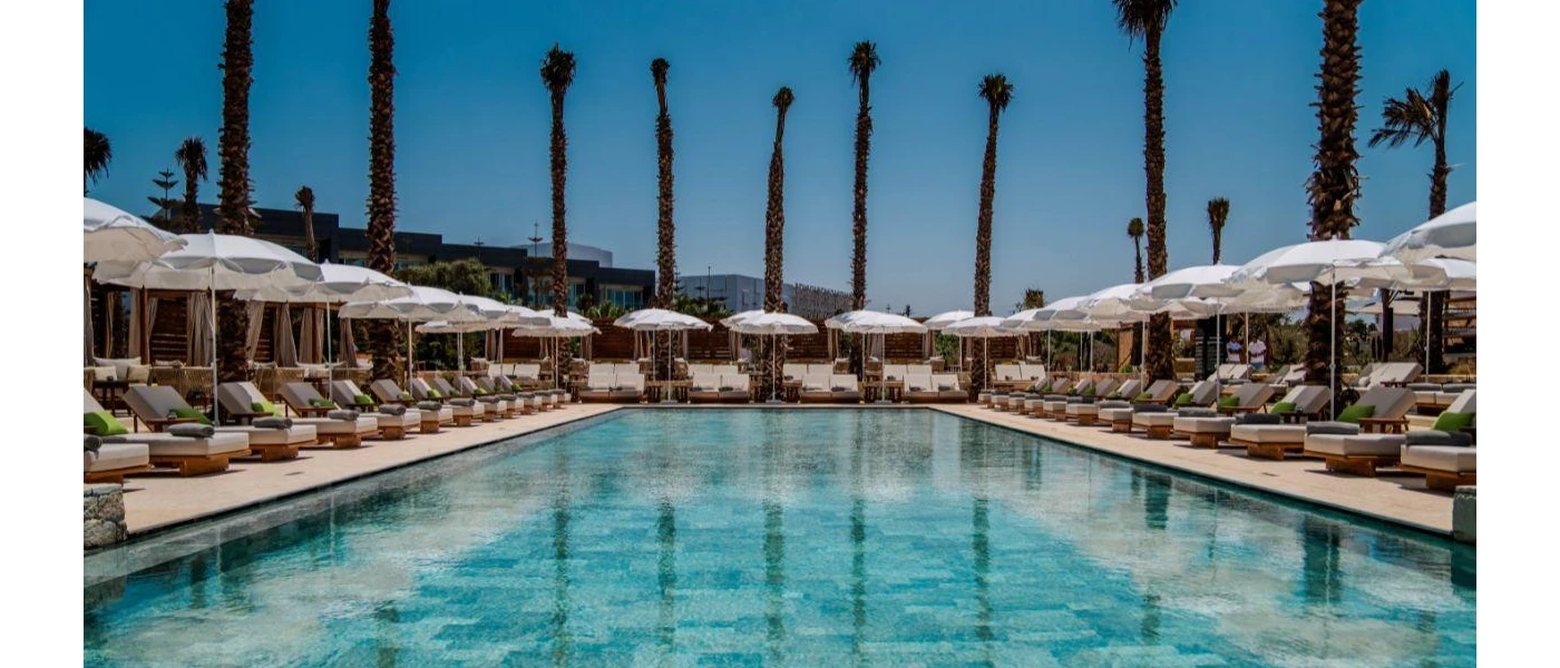Rectangular swimming pool surrounded by white umbrellas, loungers and palm trees at The View Bouznika 