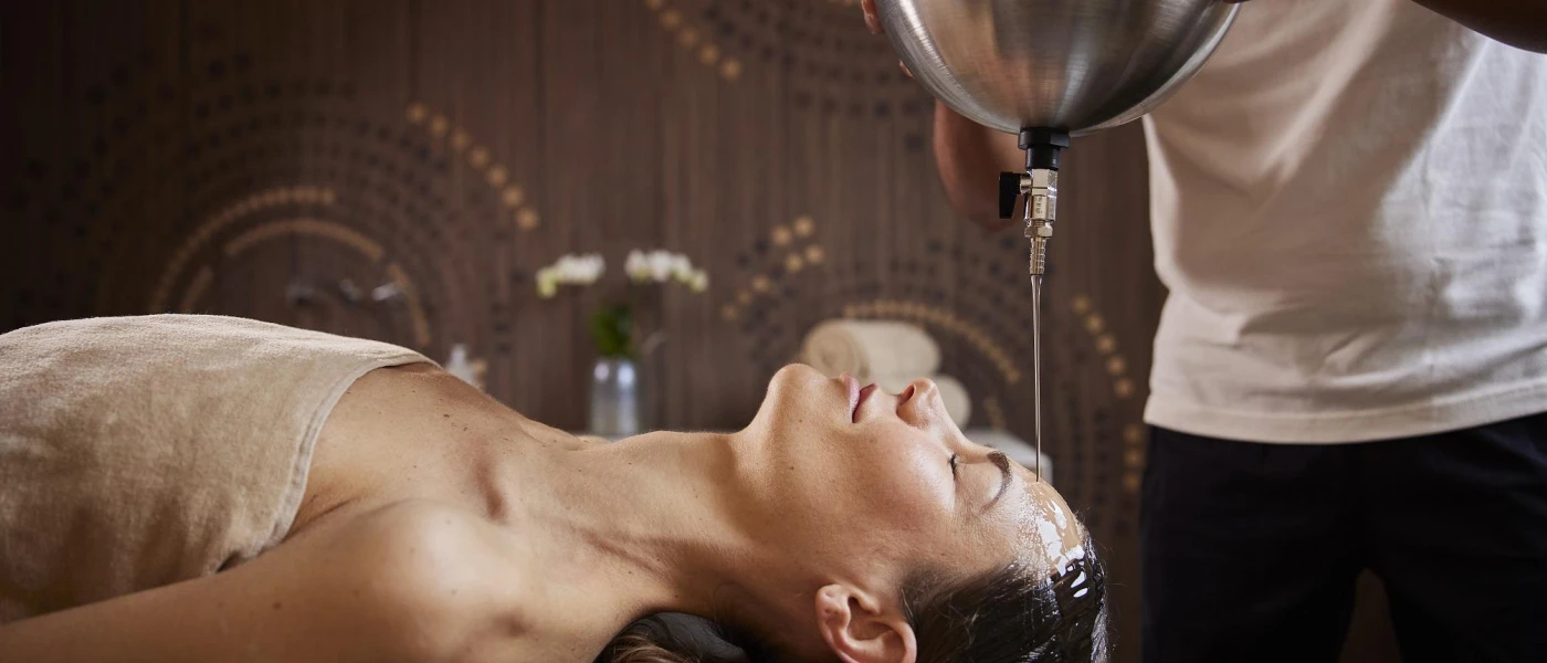 Woman lying down with her eyes closed and hair back, as a therapist pours oil over her head